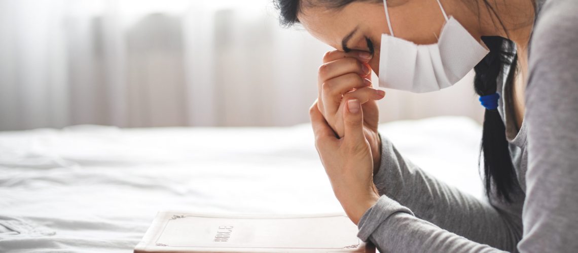 Woman with mask praying next to bed with her bible near.